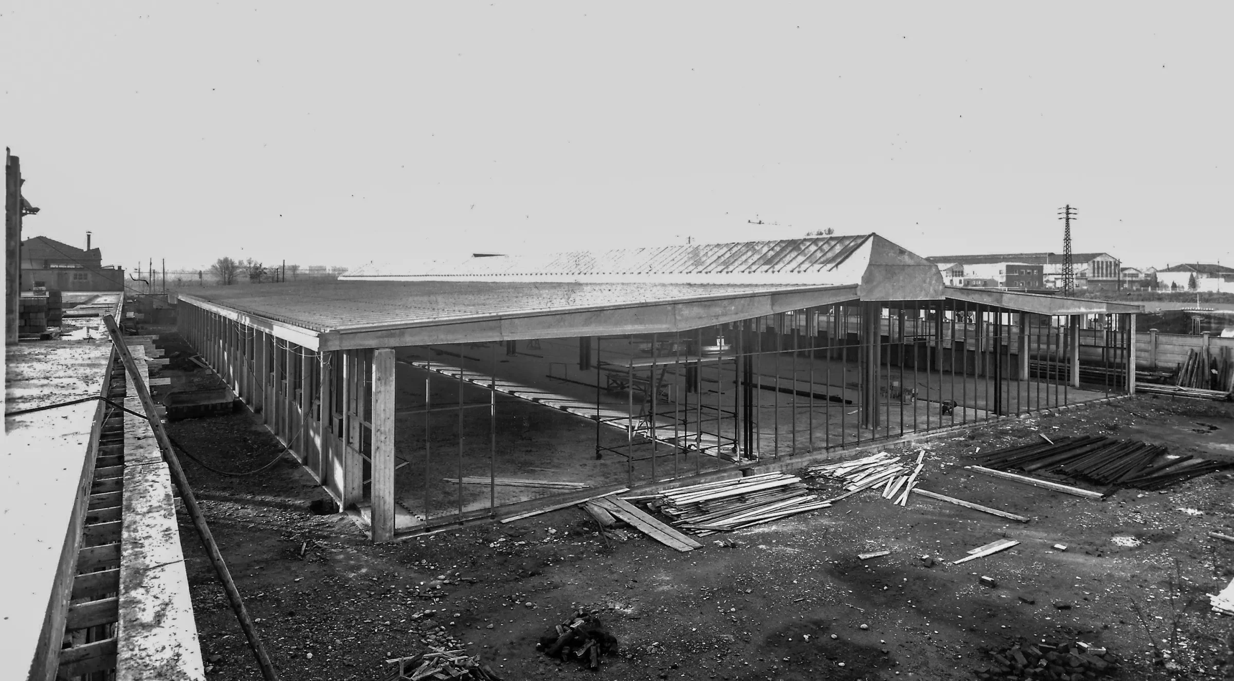 Fotografia d’archivio in bianco e nero: la struttura in cemento armato di un edificio a un piano, con la copertura già posata e il terreno del cantiere ingombro di tavole e tubi.
