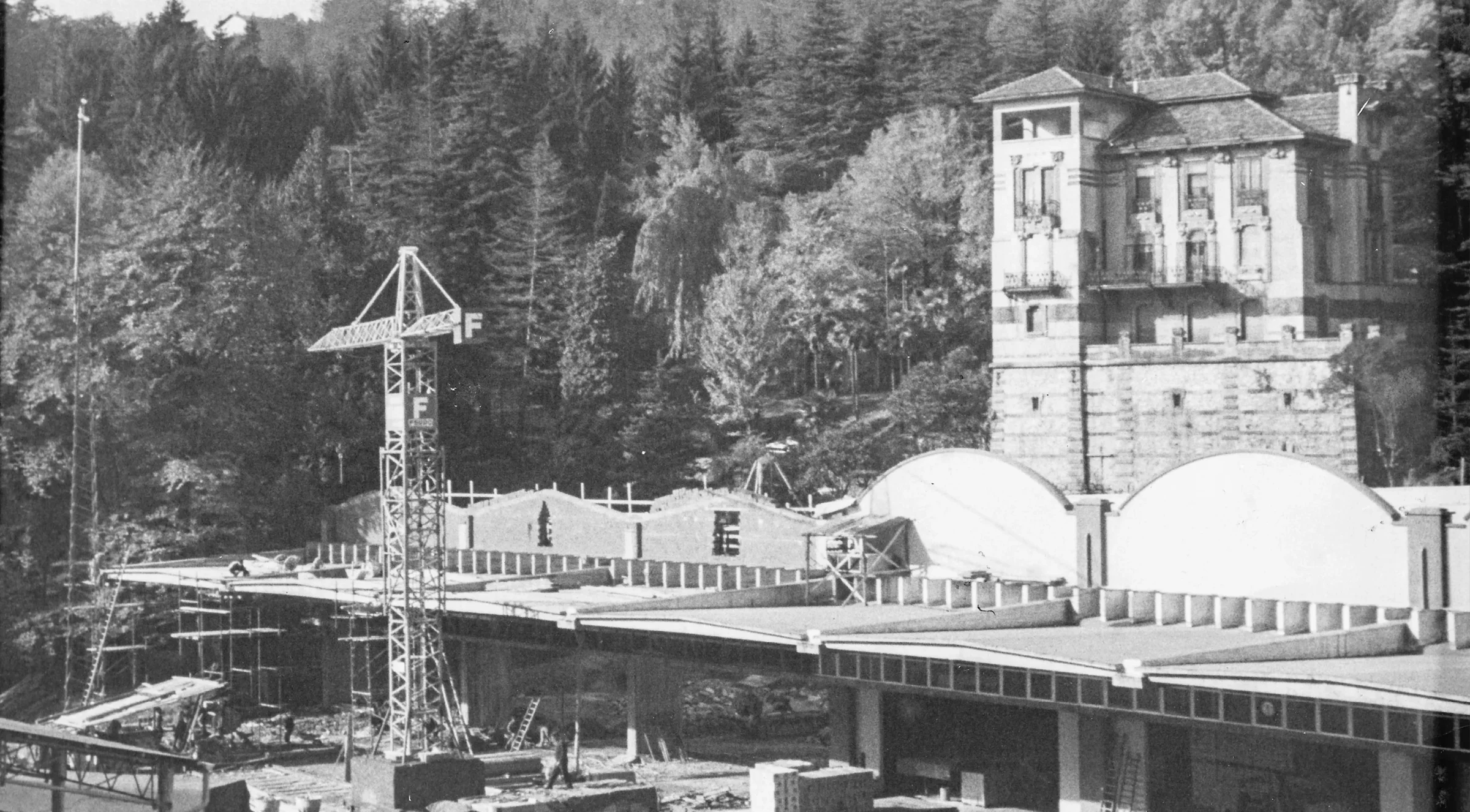 Fotografia d’archivio in bianco e nero: una gru da cantiere accanto a un edificio in costruzione ai piedi di una collina boscosa, con una villa storica a più piani sullo sfondo.